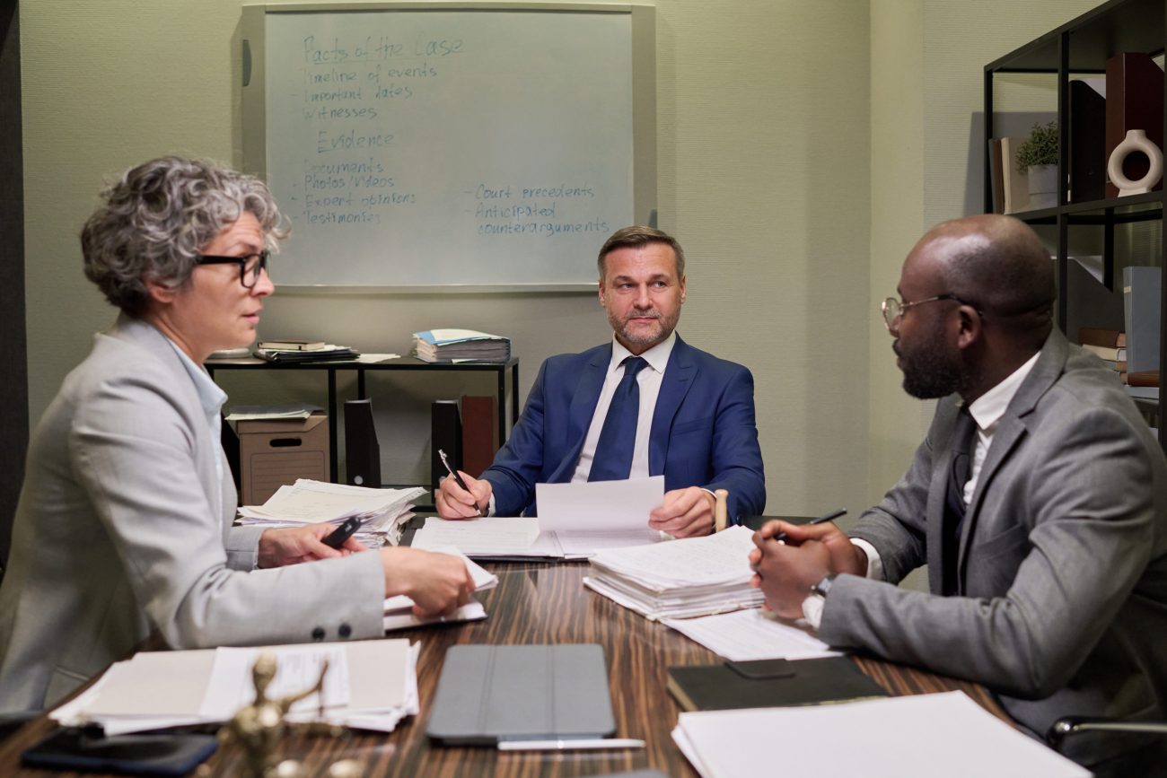 Three professionals in a meeting around a wood table with stacks of papers and a whiteboard in the background.