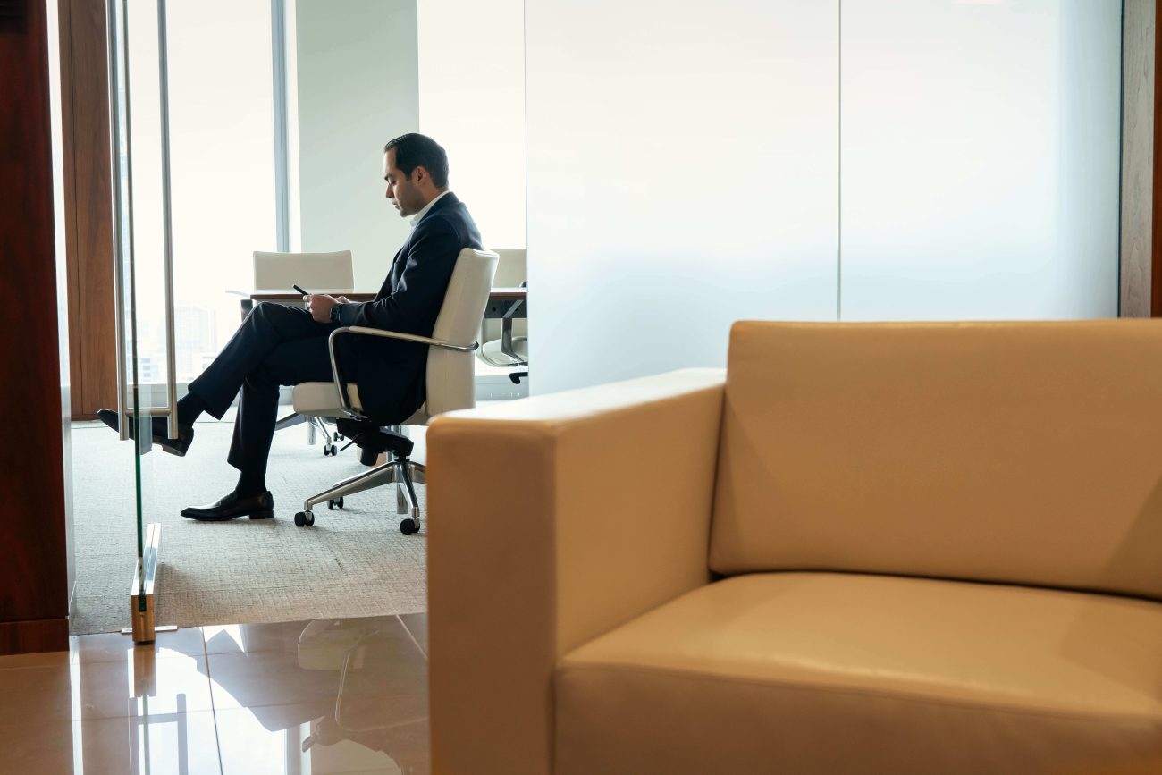 772028 - Javier Cortijo Moreno | Realidad Económica Man in a suit sits in an executive chair at a glass-walled meeting room, reading a document/tablet.