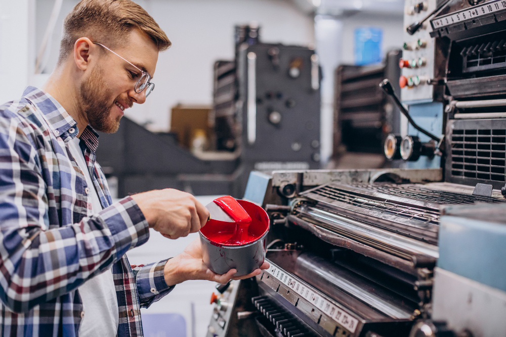 772497 - Korn Maquinaria Gráfica | Realidad Económica Man in a plaid shirt and safety glasses pours red ink into a large printing press used in a workshop.