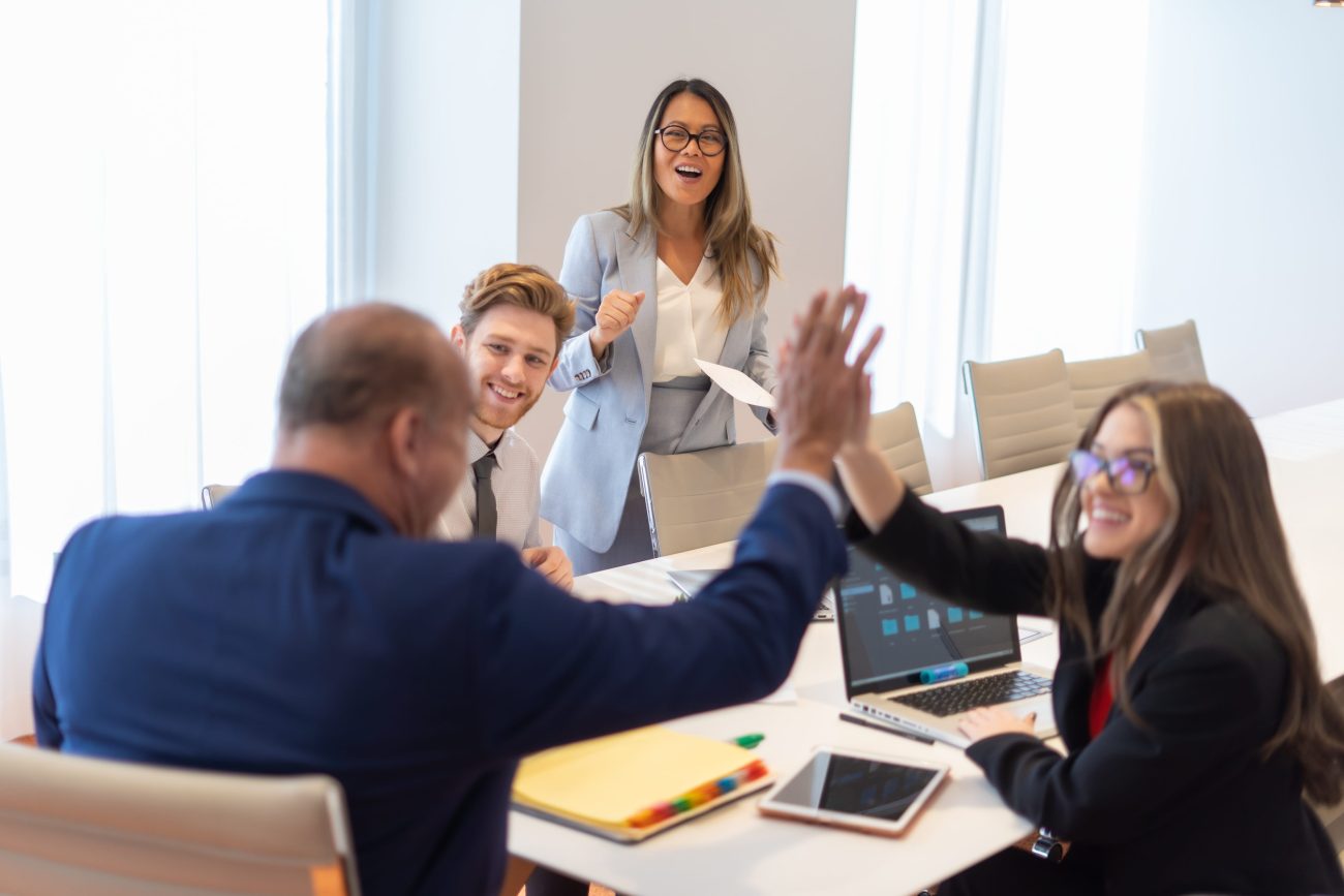Office employee having a meeting with their team | Realidad Económica Team celebrates a win with a high-five at a modern meeting table; presenter stands nearby smiling.