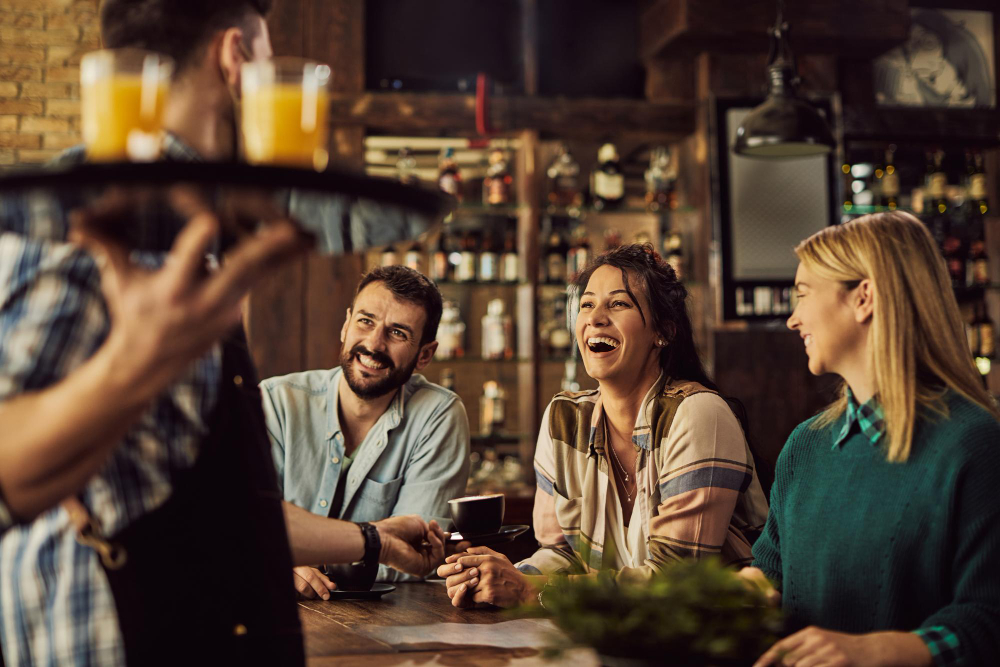 Friends laughing and chatting at a bar counter as a bartender pours drinks in the foreground.