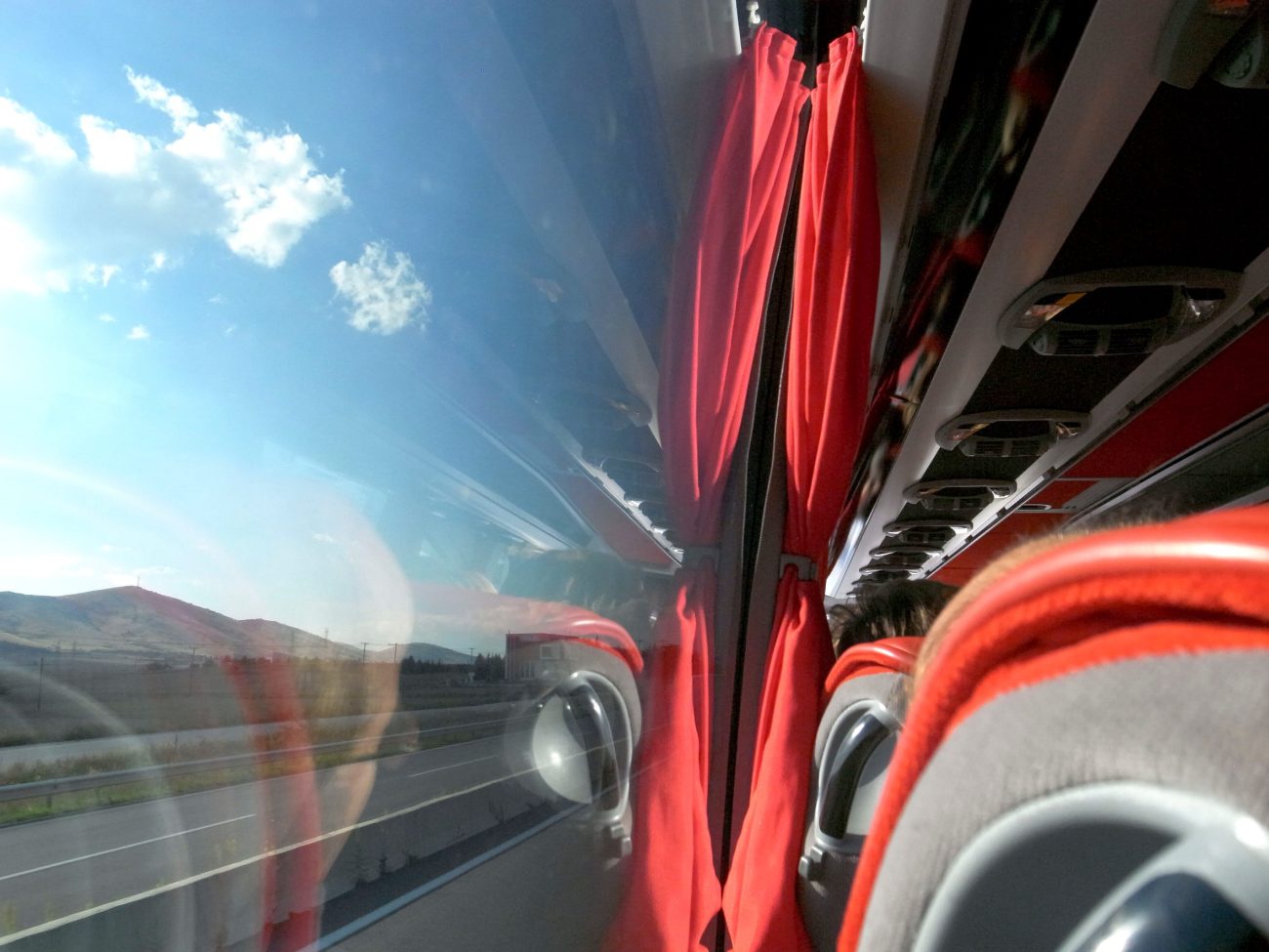 Bus interior with red curtains and gray seats, window showing highway and distant hills under a blue sky with clouds, plus a faint reflection of the photographer.