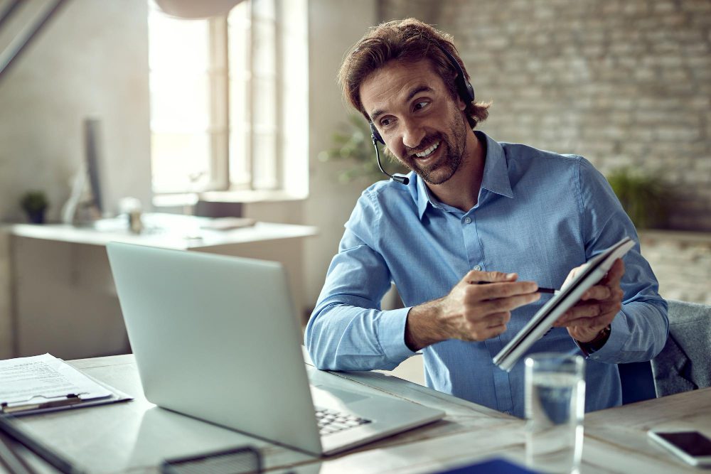 Smiling man with a headset pointing at a tablet at his desk in a bright office with a laptop nearby.