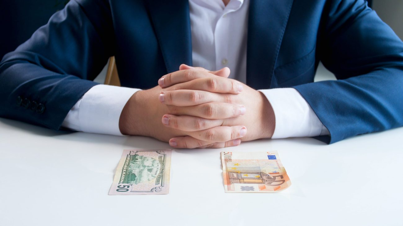 Businessperson in a blue suit with hands clasped on a white table, two banknotes (USD and EUR) laid in front.