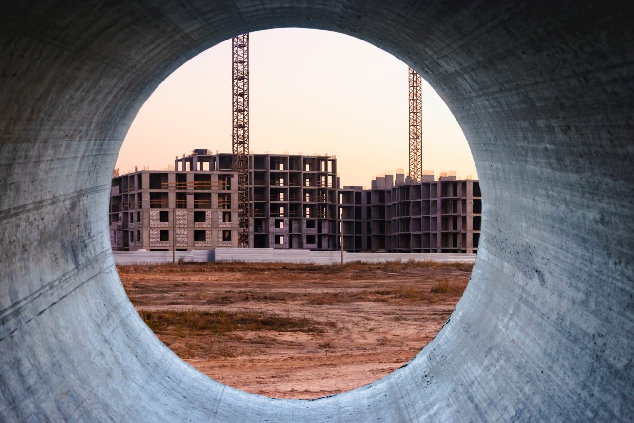 Construction site framed by a large circular concrete tunnel, showing unfinished buildings and cranes at sunset.