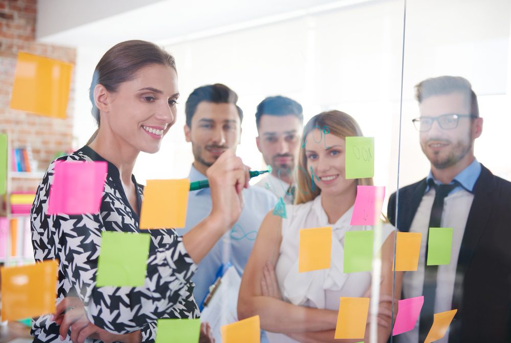 776635 - Talent Swarm | Realidad Económica Team of colleagues brainstorming on a glass wall covered with colorful sticky notes, one woman writing with a marker for ideas.
