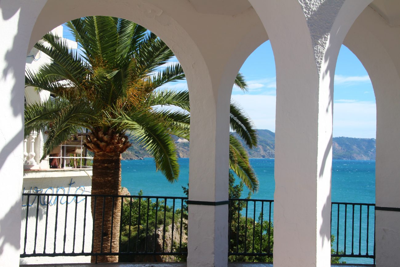 White arched colonnade framing a palm tree and the blue sea beyond.