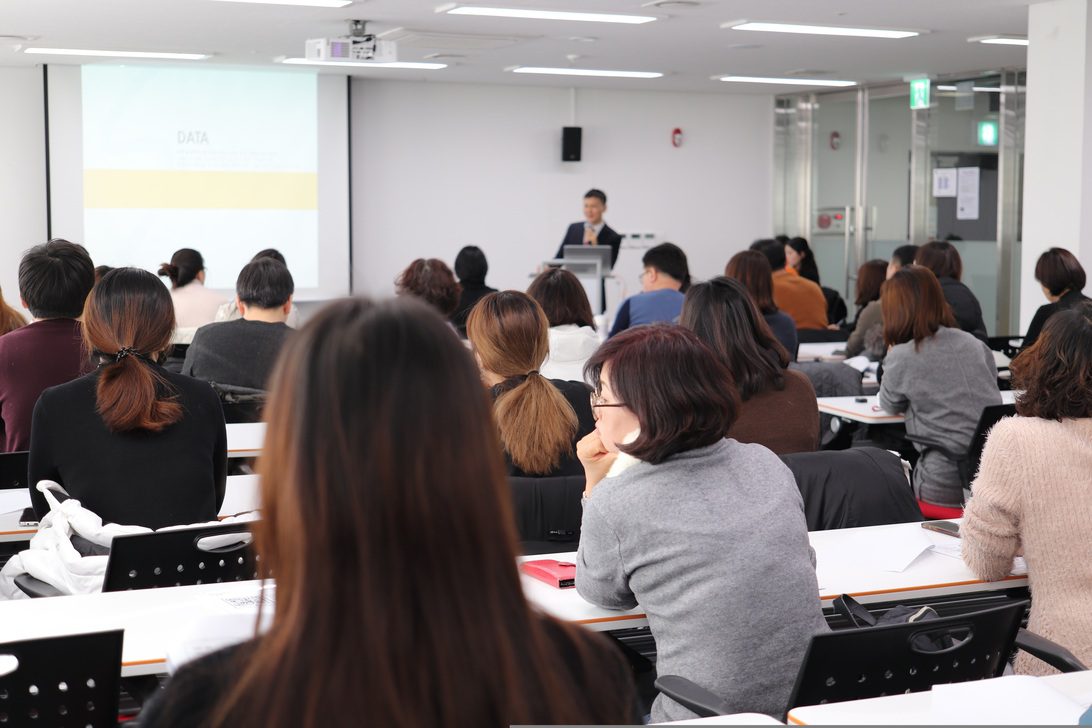 Speaker at a podium presenting to a full audience in a modern conference room.2/10