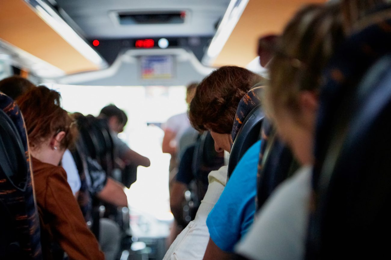 Interior of a bus with passengers seated along the aisles, viewed from the middle toward the front prior to arrival at the stop.