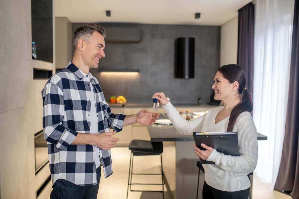 777530 - Forlares Real | Realidad Económica Man and woman in a modern kitchen exchange house keys, both smiling. The woman holds a tablet or folder while handing the keys to the man.