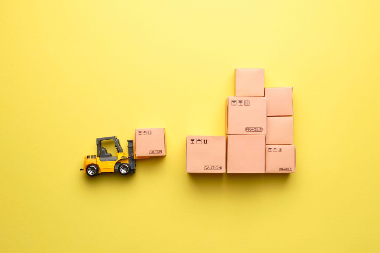 Abstract forklift for loading goods into a warehouse | Realidad Económica Miniature yellow forklift placing a cardboard box beside a stack of packed boxes on a bright yellow background.
