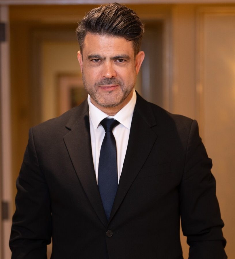 MH_PHOTOGRAPHY | Realidad Económica Man in a black suit and navy tie stands in a hallway, looking at the camera with a slight smile.