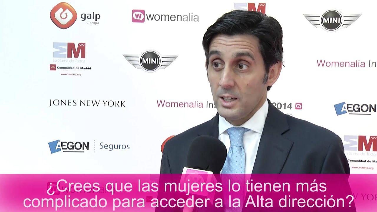 Man in a dark suit and light blue tie being interviewed with a pink caption bar in Spanish at the bottom. Logos on a white backdrop behind him indicate a media event.