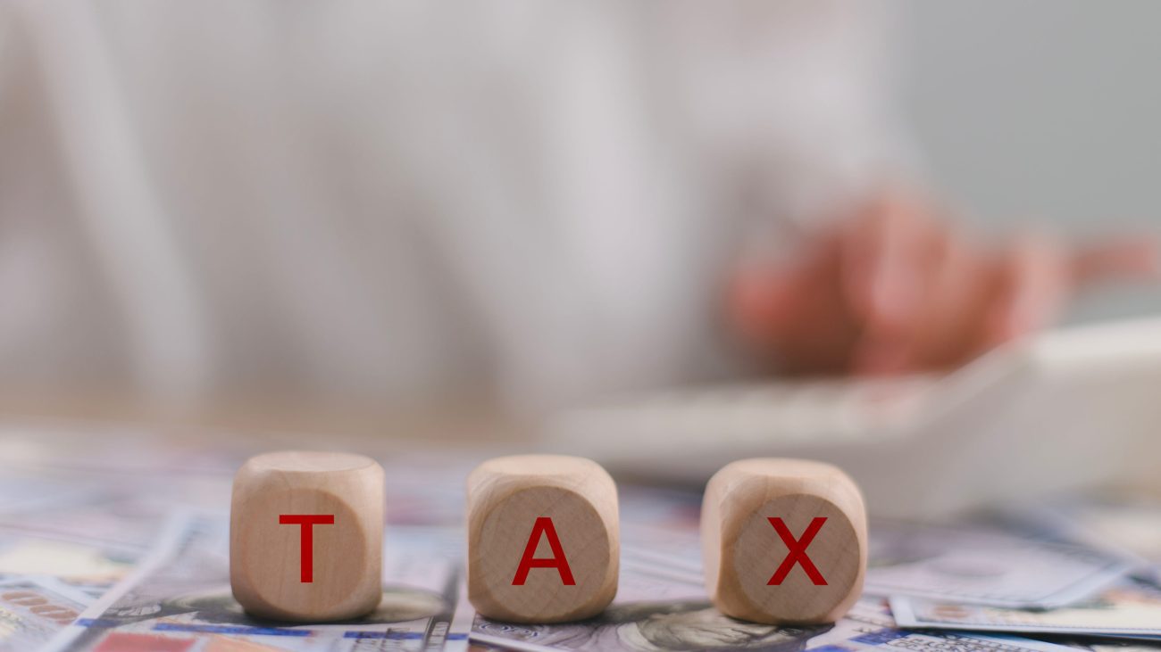 Tax word on wooden cubes with a woman working in the background | Realidad Económica Three wooden blocks spell TAX on a bed of cash, with a person typing in the background—conveys taxes or tax-related work.