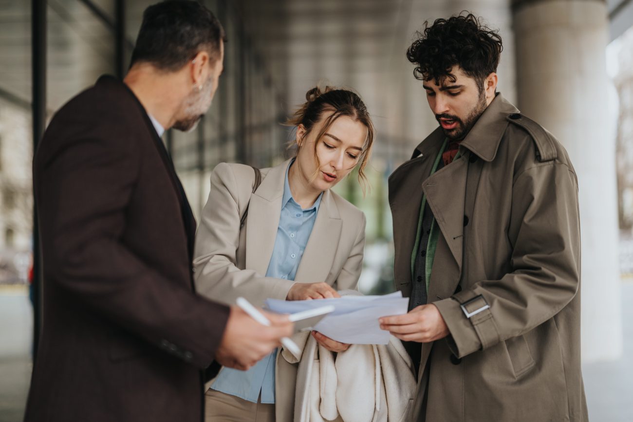 Group of colleagues discussing documents while standing outdoors | Realidad Económica Three professionals consult on documents outside a building, reviewing papers together.