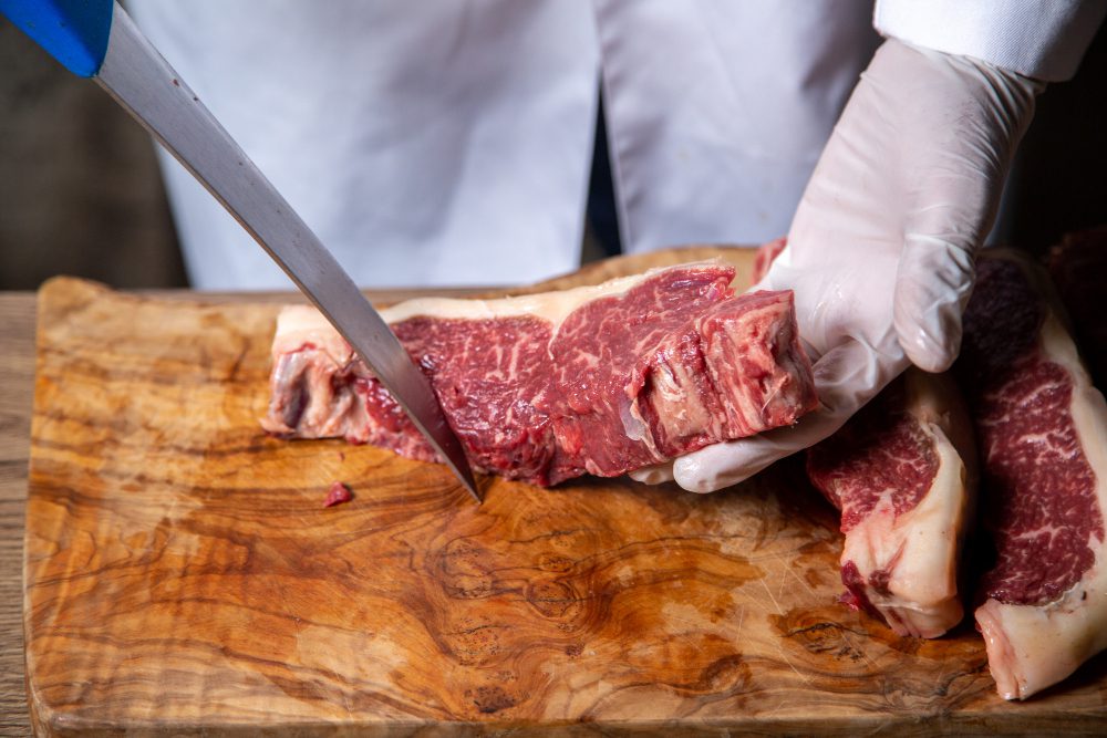 Butcher wearing gloves slices thick raw beef on a wooden cutting board with a sharp knife