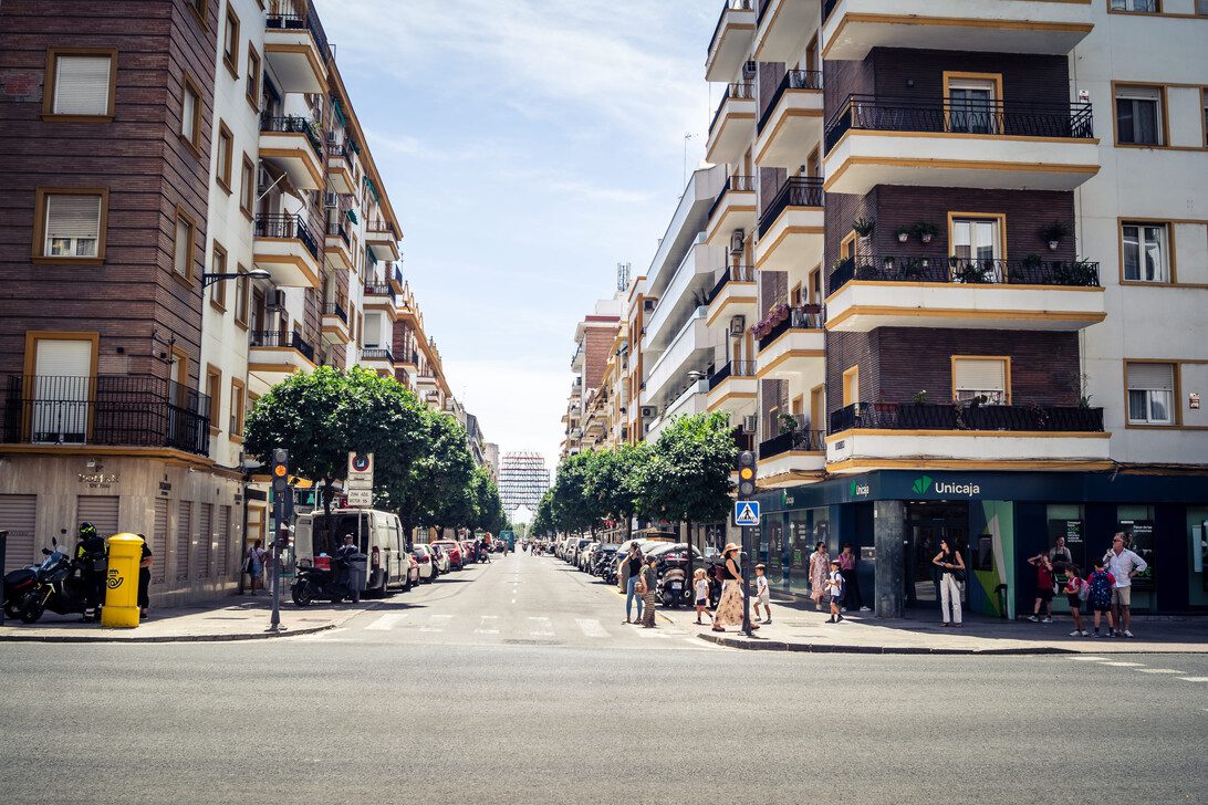 781457 Fincas Legio | Realidad Económica City street scene with mid‑rise apartment buildings, parked cars, and pedestrians crossing at a crosswalk on a sunny day.
