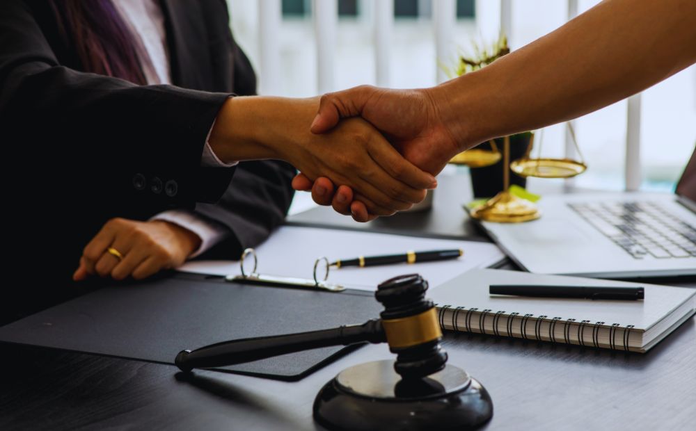 782214 - Grupo AB Abogados | Realidad Económica Two professionals shake hands across a desk with a gavel, notebook, and laptop nearby.