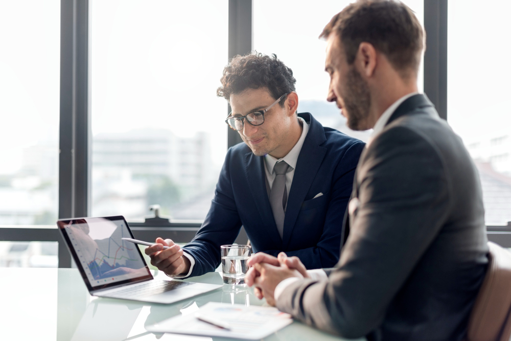 784773 - Nexum Consulting | Realidad Económica Two businessmen in suits discuss a tablet with rising graphs at a glass conference table, daylight in the background.",