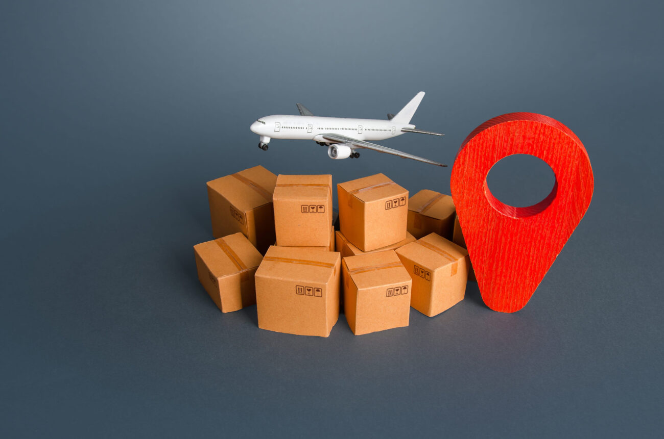 785100 IAS Handling | Realidad Económica Model airplane flies over a cluster of cardboard boxes with a large red location pin, illustrating air delivery and shipping concept.